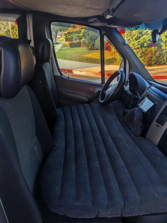 Inside an RV rental: a van fitted with a long air mattress for sleeping, seen with the dashboard and street view outside.. Mercedes Benz Sprinter 2500 2014