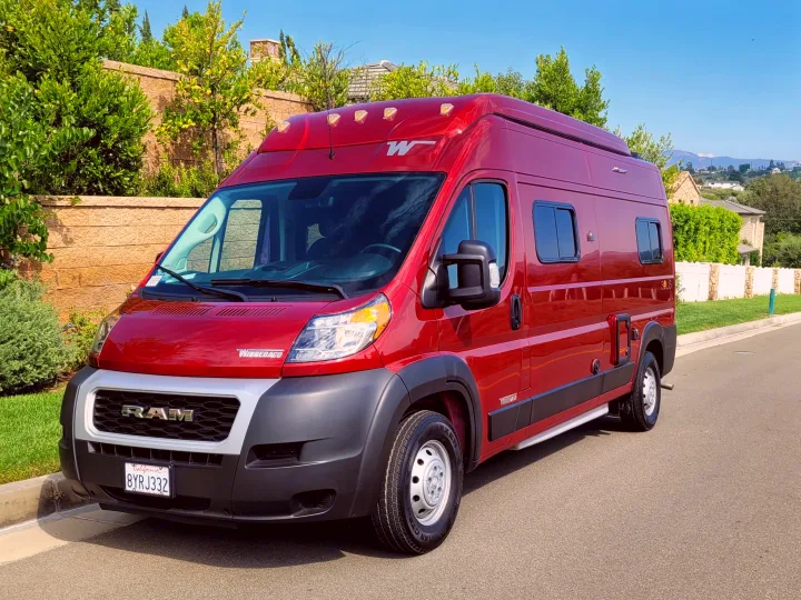 Red Winnebago Solis camper van, a perfect RV rental, parked on asphalt with green foliage and a stone wall.. Winnebago Solis 2022