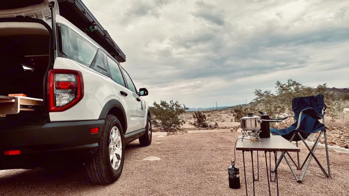 A white SUV camper with an open hatch and rooftop setup, parked in a desert. A camp stove, pot, and chair are ready. Great #rvrental.. Ford Bronco Sport 2022