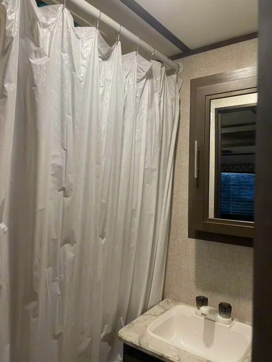 Inside an RV rental bathroom, showing a white shower curtain, sink, and mirror.. Dutchmen Coleman Lantern 2019