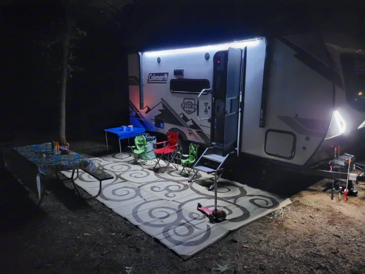 Nighttime at an RV rental campsite. A brightly lit travel trailer with an outdoor mat, chairs, and picnic table setup.. Dutchmen Coleman Rubicon 2021
