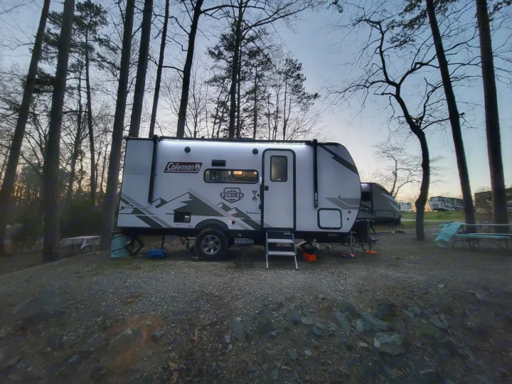 This white Coleman Rubicon RV rental is perfectly set up at a serene, tree-lined campsite. Enjoy cozy outdoor adventures at dusk.. Dutchmen Coleman Rubicon 2021