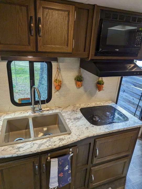 Inside an RV rental kitchen: a double sink, two-burner stove, microwave, wood cabinets, and marbled countertops.. Keystone RV Bullet Crossfire 2018