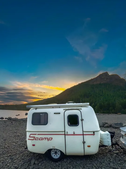 Experience the beauty of nature with an RV rental! This white Scamp camper sits by a lake at sunset, framed by a forested mountain.. Scamp Scamp Trailer 2007