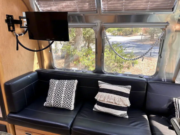 Inside an RV rental, a black leather sofa with patterned pillows faces a mounted TV. Large windows offer a view of trees and a road.. Airstream Flying Cloud 2016