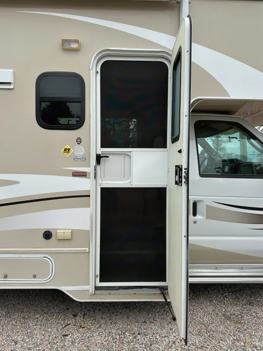 RV rental: Side view of a tan Winnebago motorhome's open entry door with screen, parked on gravel.. Winnebago Minnie Winnie 2014