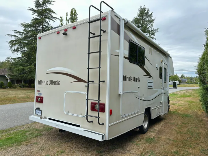 A beige Minnie Winnie RV rental is parked on grass, displaying its rear, side, ladder, and awning under a cloudy sky.. Winnebago Minnie Winnie 2014