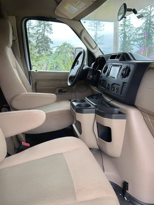 Spacious front cabin of an RV rental, showcasing light-colored seats, steering wheel, dashboard, and a view of trees.. Winnebago Minnie Winnie 2014