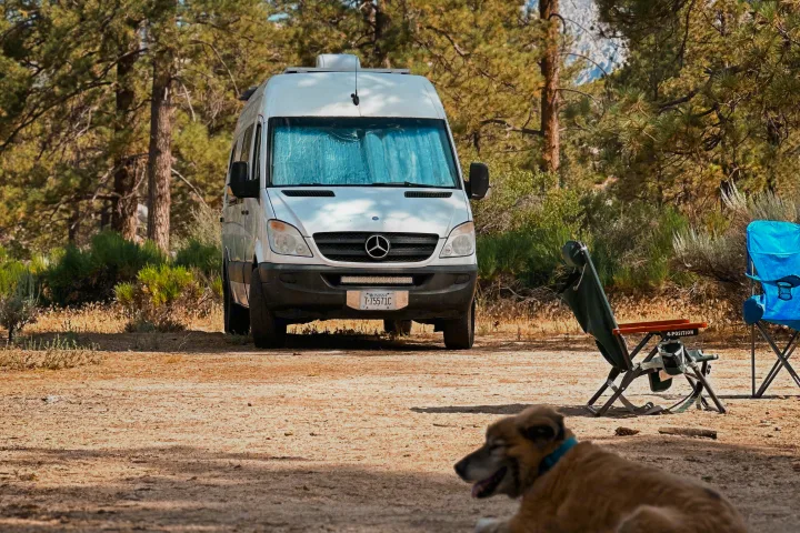 A white Sprinter RV rental parked at a serene campsite with two chairs and a dog enjoying the outdoors among pine trees.. Mercedes-Benz Sprinter 2013