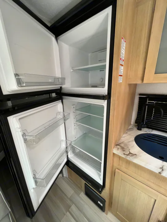 Inside an RV rental: a black, two-door refrigerator stands open, revealing its clear shelves, alongside a kitchen sink.. Forest River Salem FSX 2023