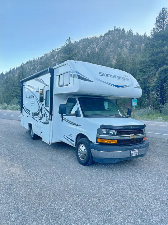 A white Sunseeker RV rental is parked on a gravel road shoulder, with scenic forested mountains and clear blue sky behind it.. Forest River Sunseeker LE 2020
