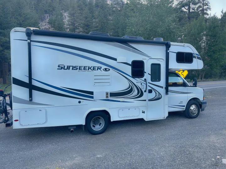 This Sunseeker RV rental motorhome, white with blue graphics and a bike rack, is parked on a gravel road amid trees.. Forest River Sunseeker LE 2020