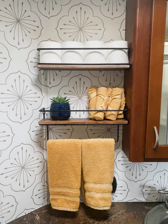 Organized RV rental bathroom. Wall shelves hold toilet paper, yellow towels, and a plant, offering smart storage in a compact space.. Forest River Vengeance 2015
