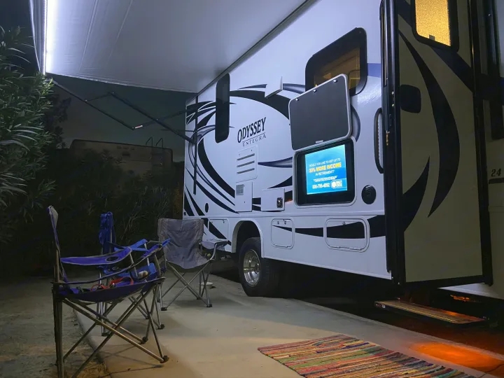 RV rental at night: Bright awning, outdoor TV for entertainment, and chairs set up for a relaxing evening at the campsite.. Entegra Coach Odyssey 2020
