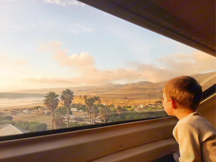 Golden hour view: A child looks out to a beach, ocean, palm trees, and an RV park. Find your ideal RV rental getaway!. Entegra Coach Odyssey 2020