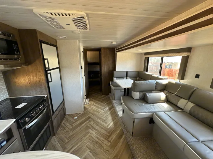 A modern rv rental interior with a kitchen, large gray sectional sofa, dinette table, and herringbone wood-look flooring.. Forest River Wildwood X-Lite 2022
