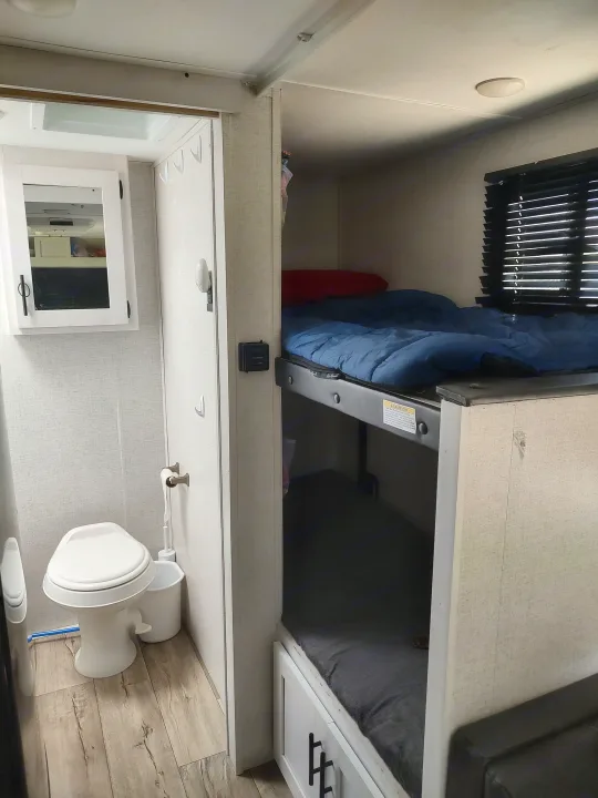 RV rental interior showing a narrow bathroom with a toilet on the left and a cozy set of bunk beds on the right.. Dutchmen Coleman Rubicon 2021