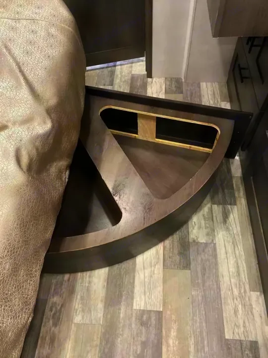 Close-up of an RV rental bed base, revealing a curved, built-in storage compartment. Gray wood-look flooring.. Forest River 34DS 2022