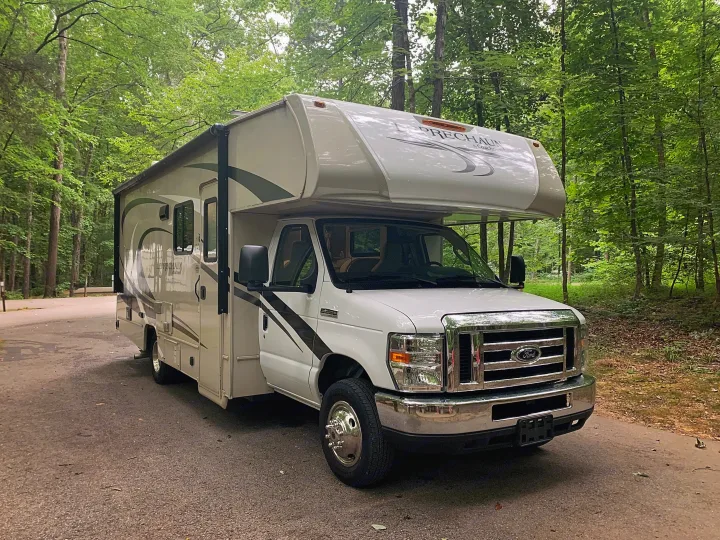 White Coachmen Leprechaun RV rental parked on a quiet campground road amidst lush green trees.. Forest River Coachmen Leprechaun 2020