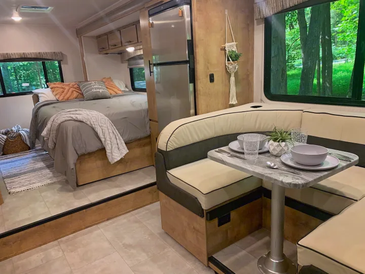 This inviting rv rental features a cozy bedroom with a fluffy bed, and a dining area set for a meal, with green trees outside.. Forest River Coachmen Leprechaun 2020