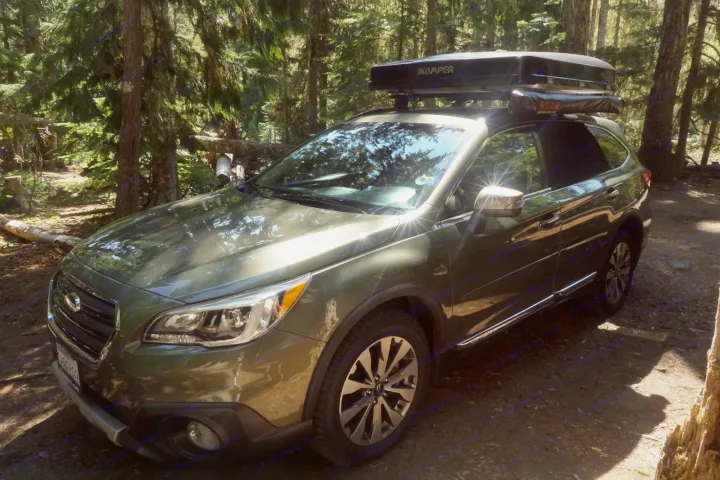 Green Subaru Outback with rooftop tent, ideal RV rental for forest camping. Parked on dirt under sunny trees.. Subaru Outback with iKamper 2017