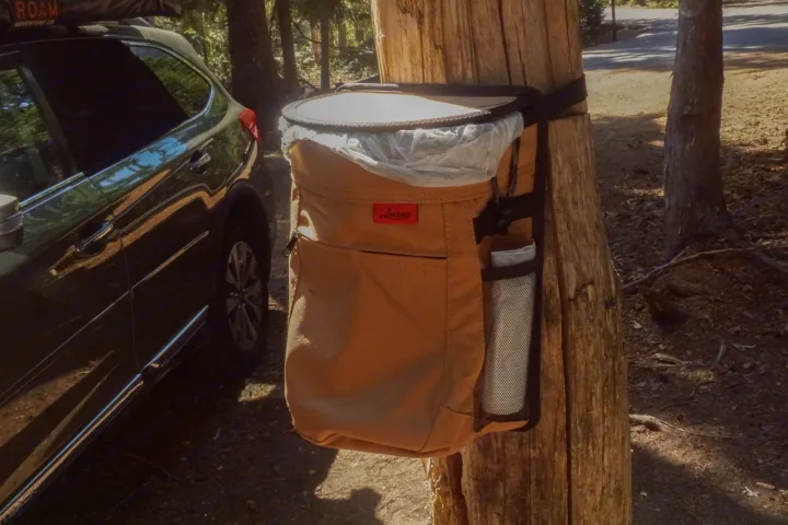 Practical campsite setup for an RV rental trip! A portable trash can hangs on a tree next to an SUV, ready for outdoor living.. Subaru Outback with iKamper 2017