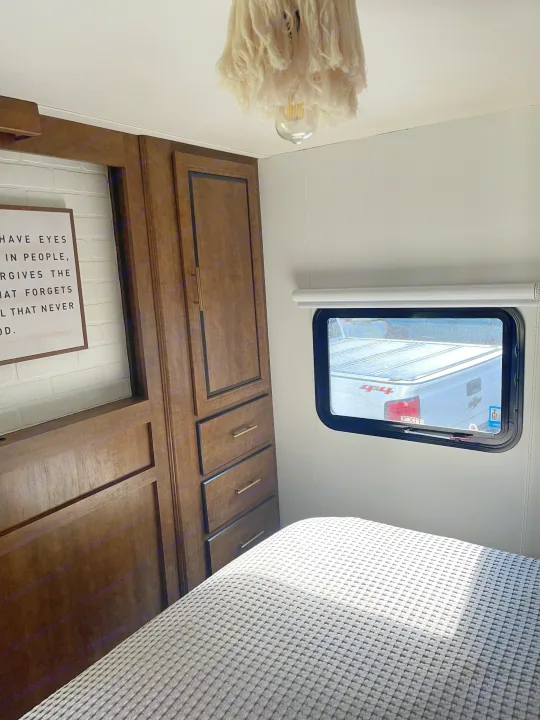 Comfortable RV bedroom showing a grey patterned bed, wood cabinets, and a small window overlooking a truck. Ideal RV rental space.. Crossroads RV Zinger 2017