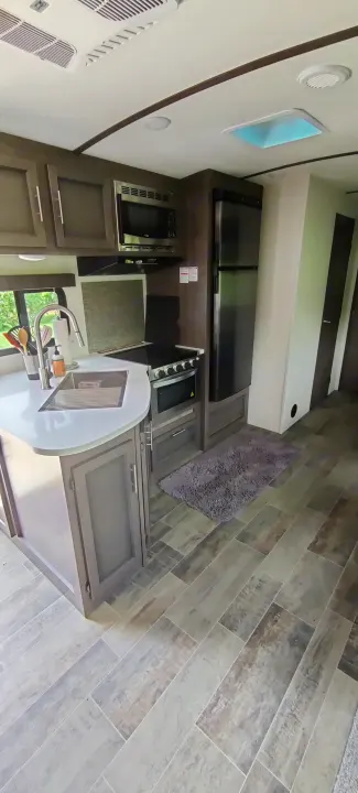 Modern RV rental kitchen with light counters, sink, microwave, stove, and fridge. Features gray cabinets, wood-look floor, and ceiling skylight.. Crossroads RV Sunset Trail Super Lite 2021