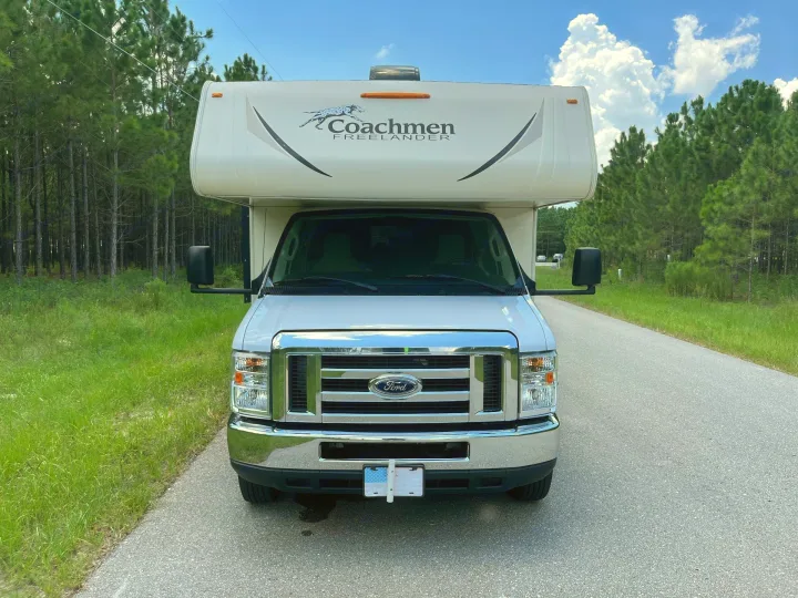 Front view of a white Ford Coachmen Freelander Class C RV rental on a road, flanked by green grass and pine trees.. Forest River Coachmen Freelander 2018