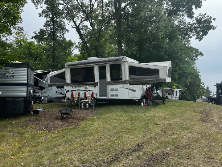 White pop-up camper RV rental at a grassy, treed campground with a fire pit and chairs. Other RVs are visible in the background for camping.. Forest River Rockwood Premier 2009