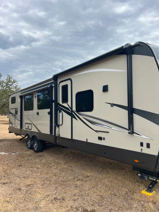 Tan RV rental, a large travel trailer, stands on dry grass under a cloudy sky. Side view showing windows and an awning.. Forest River Flagstaff Classic 2022