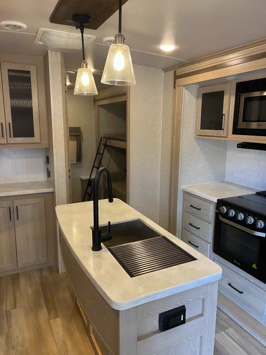 Modern RV rental kitchen featuring an island with sink, oven, microwave, light wood cabinets, and bunk beds in the background.. Forest River Flagstaff Classic 2022