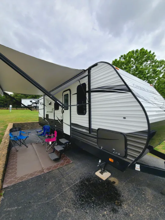 RV rental set up at a campsite with its awning extended, showing outdoor chairs and a mat for relaxation.. Highland Ridge RV Open Range Conventional OT26BHS 2019