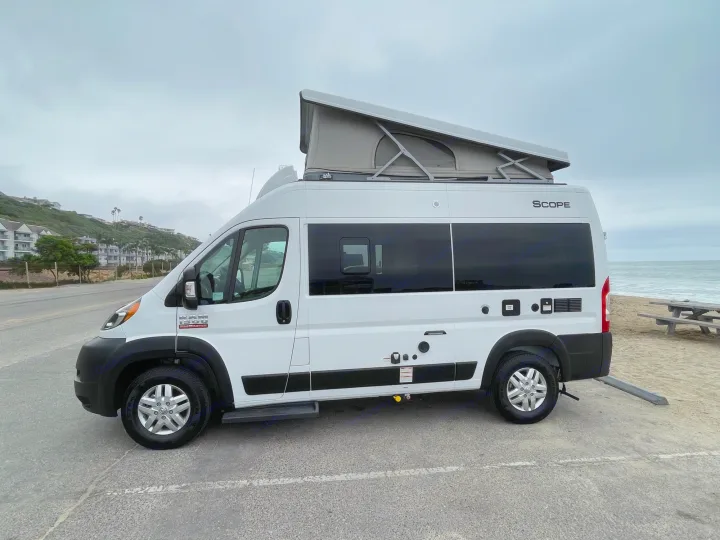 White pop-top RV rental, a camper van, parked on pavement overlooking a sandy beach and ocean.. Thor Scope 2023