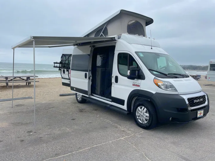 White pop-top camper van, an RV rental with an extended awning, ready for beach camping by the ocean.. Thor Scope 2023