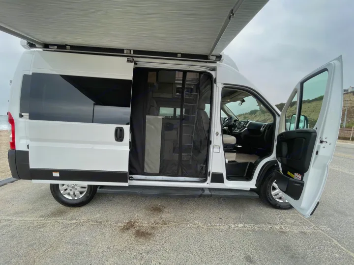 White camper van RV rental, with awning out & doors open to its interior, parked by the ocean on a cloudy day.. Thor Scope 2023