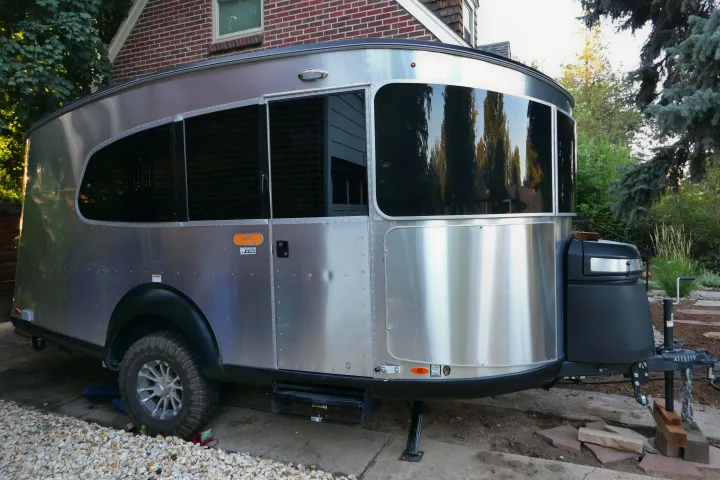 A sleek silver RV rental trailer is parked beside a brick house. Large tinted windows reflect trees.. Airstream Basecamp 2023