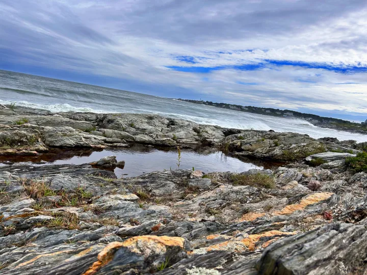 Low-angle view of rugged rocks surrounding a still pool, ocean waves beyond, and distant coastal homes. Plan your trip with an RV rental.. Winnebago Revel 2022