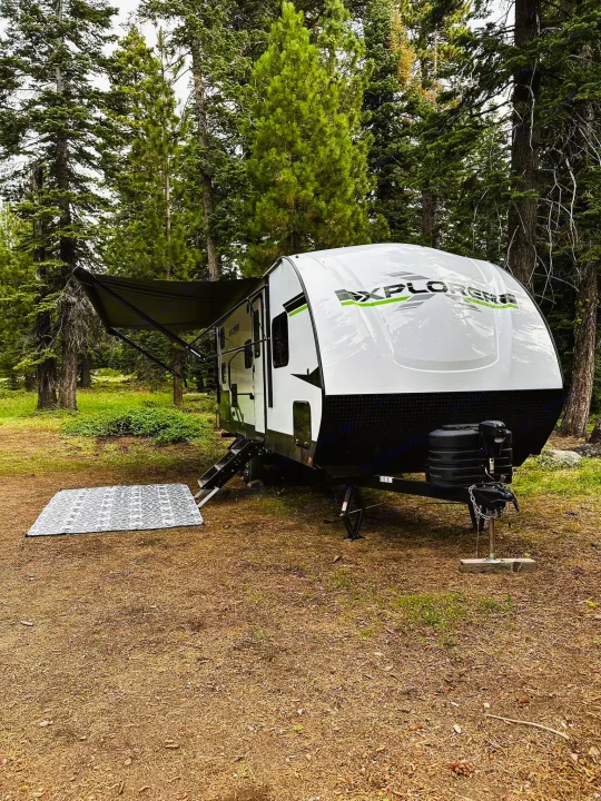 White RV rental travel trailer with awning extended, set up in a pine forest campsite with a mat. Ready for your camping adventure!. Riverside RV Xplorer 240BHx 2023