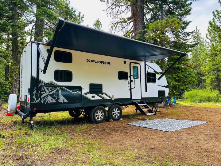 An Xplorer RV rental, white with dark accents, is perfectly set up at a serene forest campsite with its awning extended.. Riverside RV Xplorer 240BHx 2023
