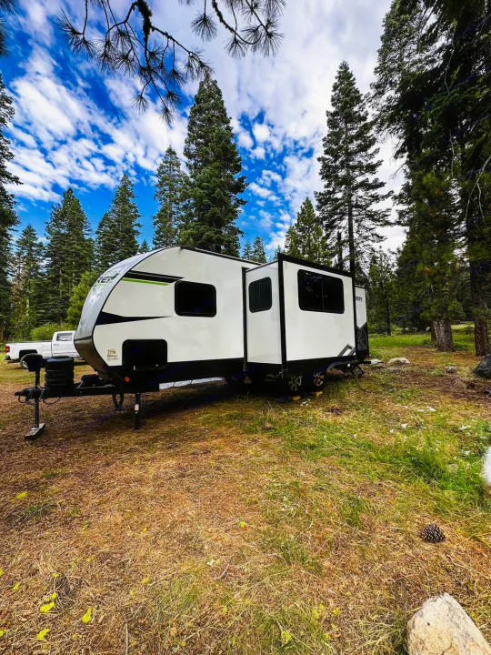 Enjoy nature! This RV rental is set up for camping in a serene forest, with tall trees under a vibrant sky.. Riverside RV Xplorer 240BHx 2023