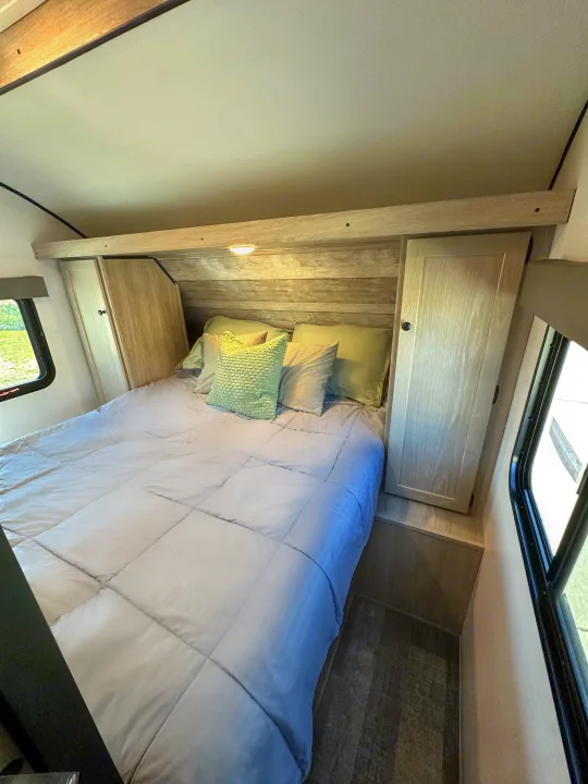 Cozy RV rental bedroom with a light gray bed, accent pillows, wooden headboard, side cabinets, and window view.. Riverside RV Xplorer 240BHx 2023