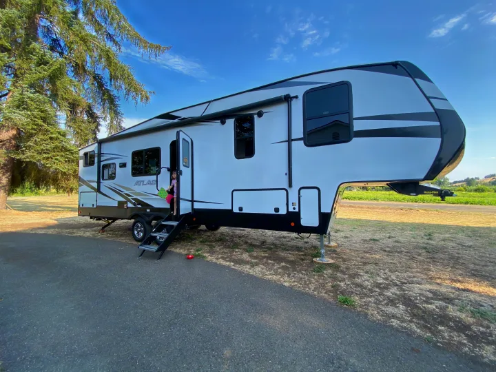 Experience an RV rental: a modern fifth-wheel parked at a sunny campsite, a child peeking from the door.. Atlas Atlas Fifth Wheel 2020