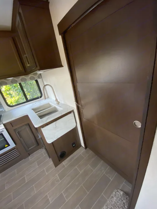 Compact RV rental kitchen with sink, marble-look counter, and dark wood cabinets, beside a space-saving sliding door.. Atlas Atlas Fifth Wheel 2020