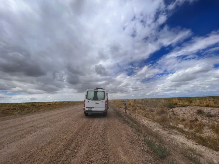 White RV rental van on a remote dirt road in a wide-open, dry landscape, under a partially cloudy sky. A person stands beside it.. Mercedes-Benz Sprinter 2012