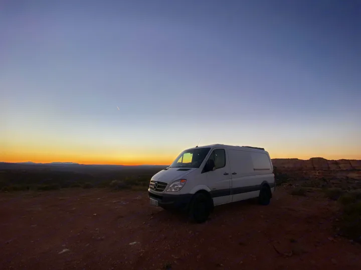 Experience van life in an RV rental Sprinter van. Parked on a dirt road, this white camper van enjoys a vibrant desert sunset.. Mercedes-Benz Sprinter 2012