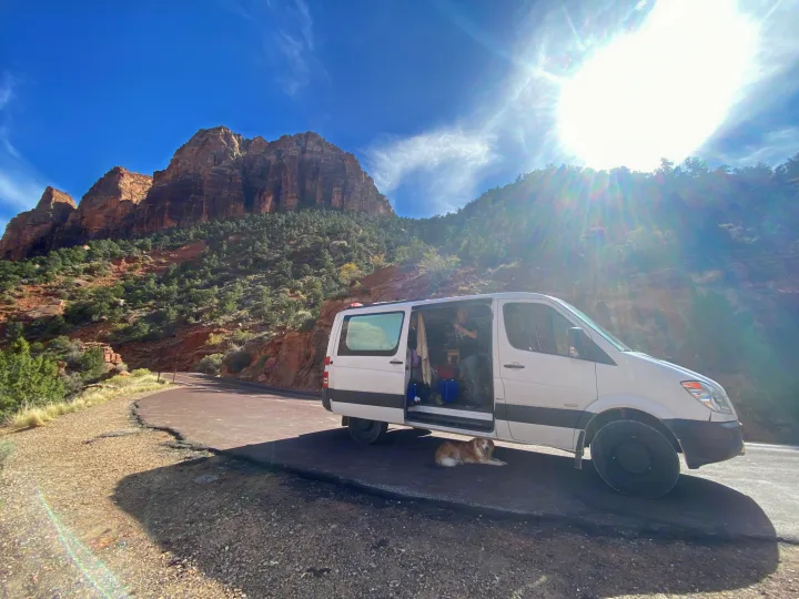 White van (likely RV rental) parked by majestic red rock mountains. A person is inside, and a dog rests under the van.. Mercedes-Benz Sprinter 2012