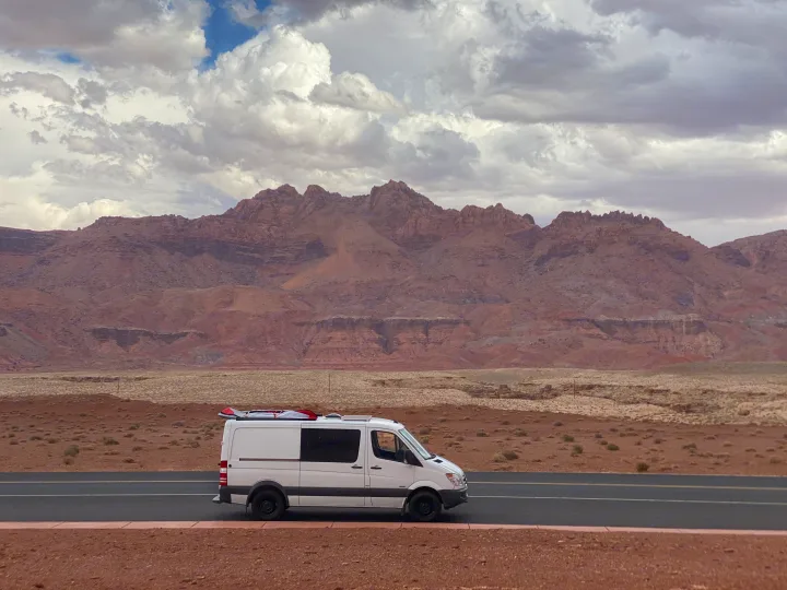White camper van (possibly RV rental) parked by a desert road, with majestic red mountains under cloudy skies.. Mercedes-Benz Sprinter 2012