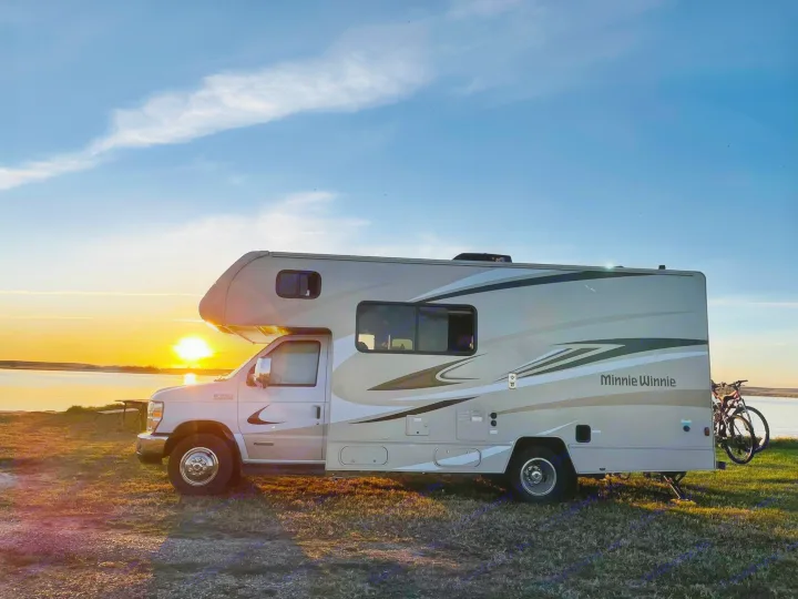 A beige Minnie Winnie RV rental is parked by a lake at sunset, with bikes on the back. Golden light bathes the scene.. Winnebago Minnie Winnie 2020