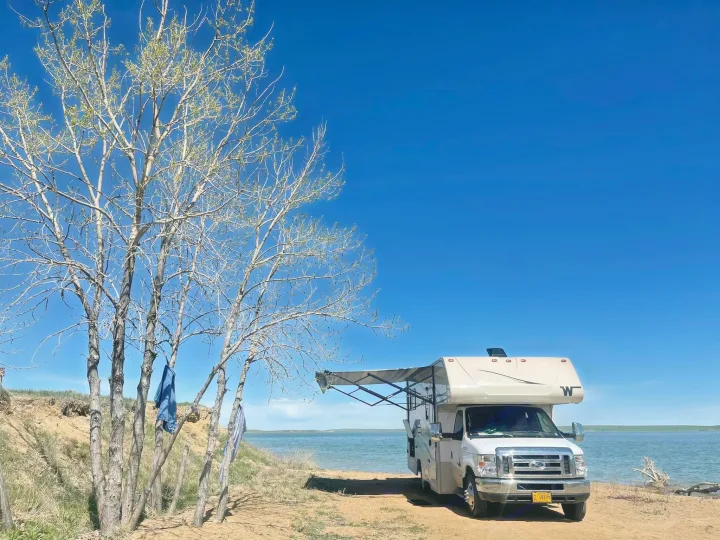 Lakeside RV rental adventure: A white motorhome with an extended awning parked by a lake, with trees and a clear blue sky.. Winnebago Minnie Winnie 2020